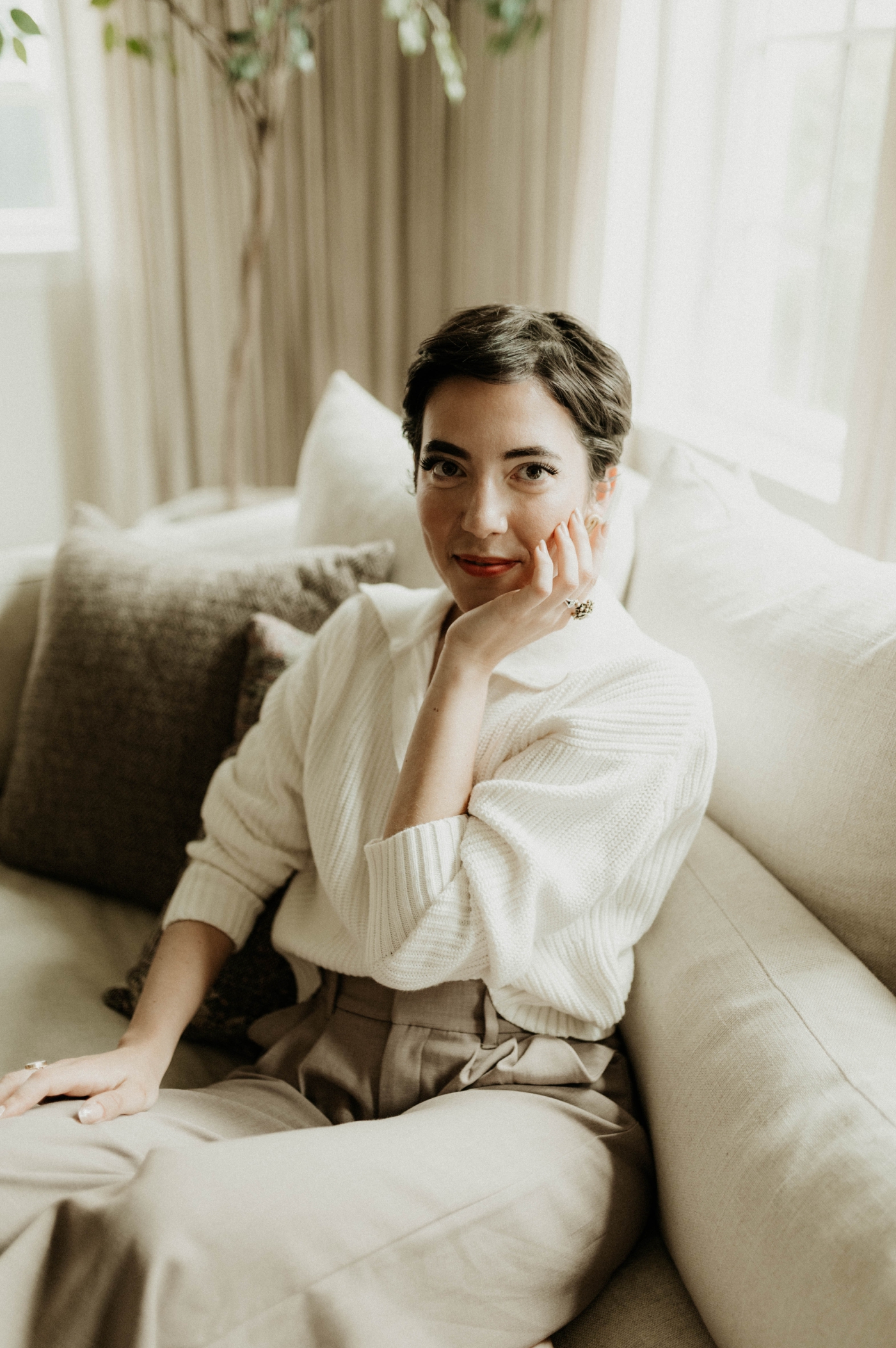 A woman with dark hair in a pixie cut sits on a white couch and smiles at the camera